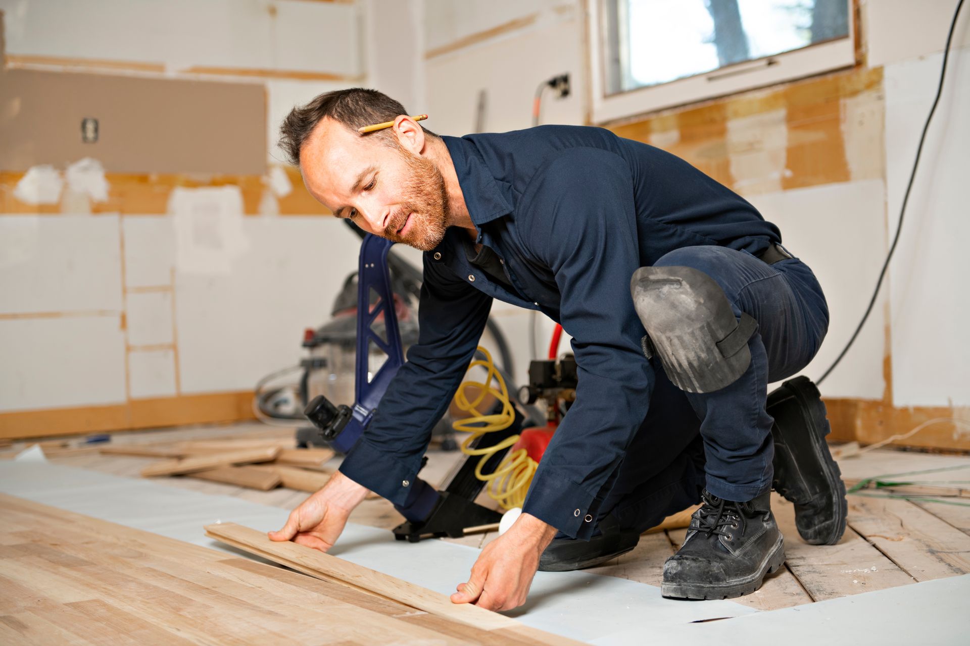 Man installing hardwood flooring, kneeling, wearing knee pads and a blue jumpsuit, indoors.