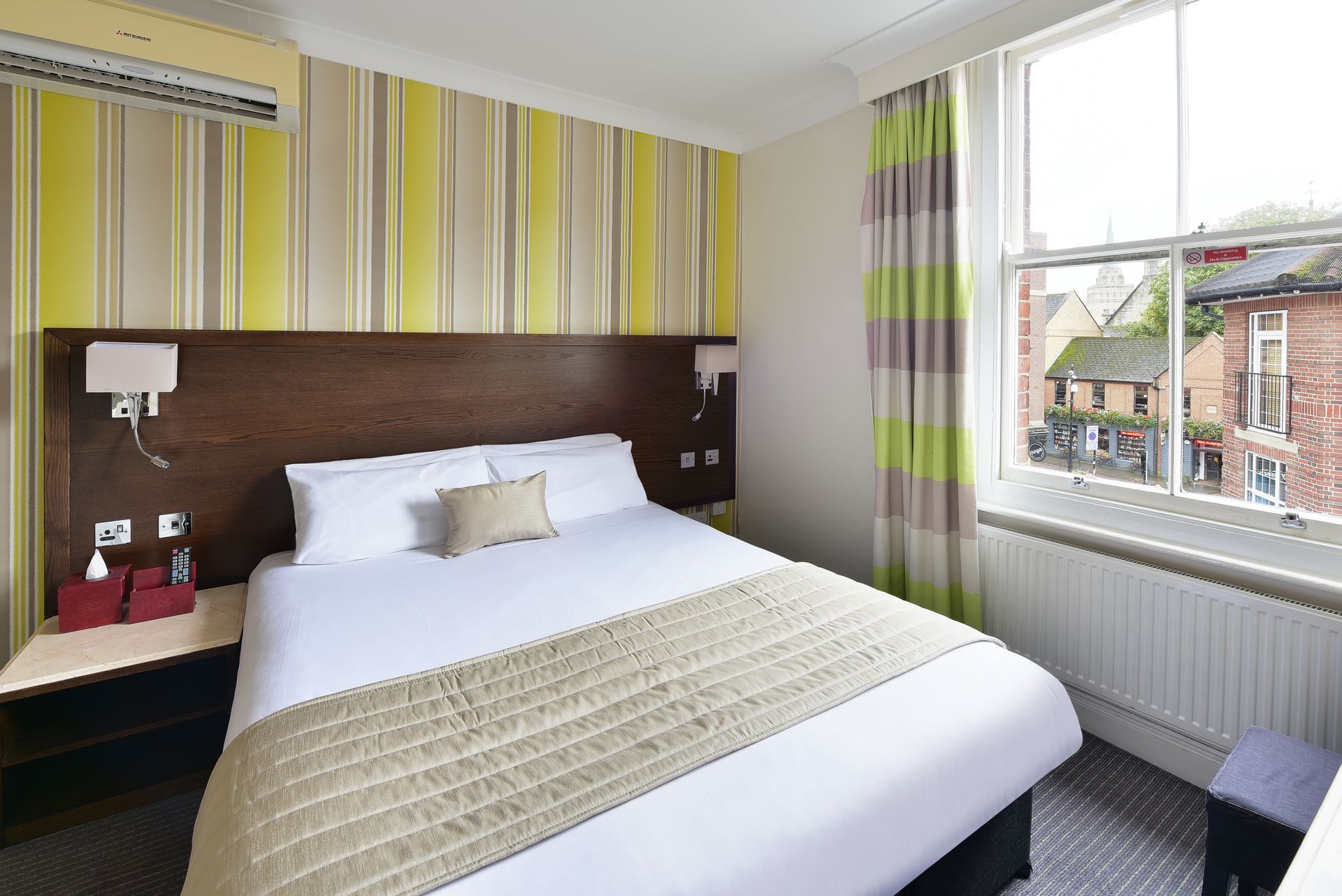 A tidy hotel room with a queen bed, yellow striped accent wall, and a large window looking out onto a street.