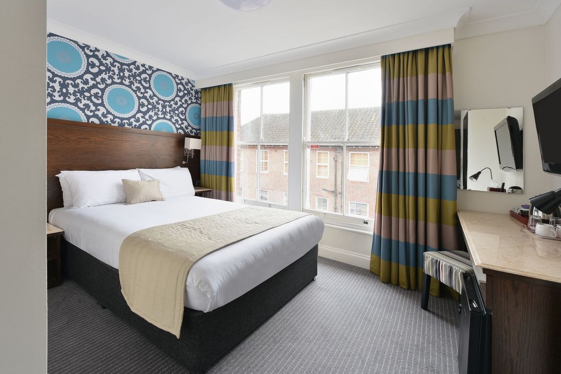 A hotel room with a queen bed, patterned blue wallpaper, striped curtains, a desk, and a mirror.