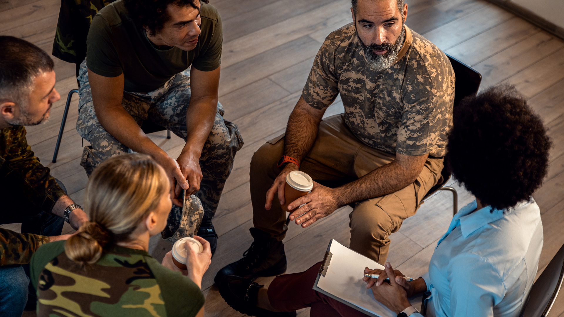 Group of people in a circle, some wearing military attire, talking with a counselor indoors.