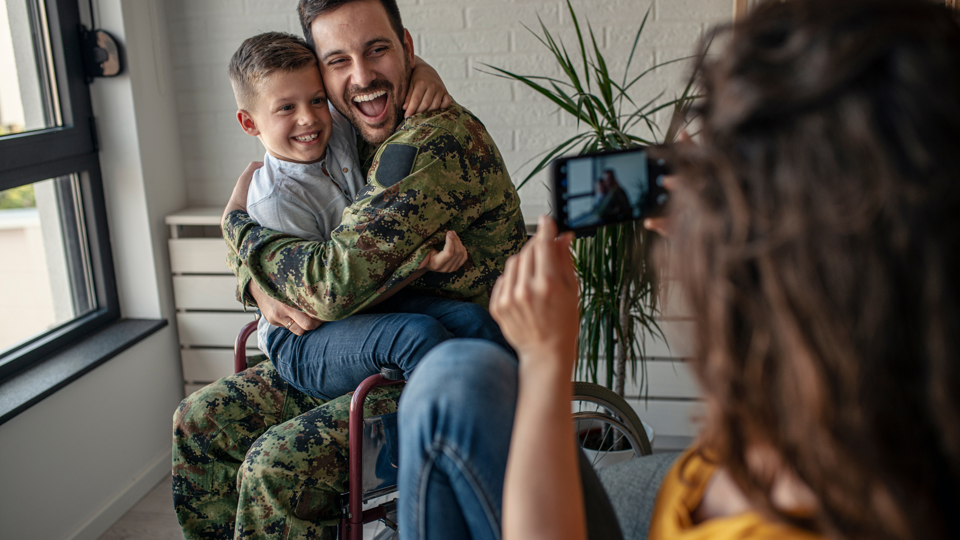 Soldier in uniform hugs a child; a woman takes their photo with a phone.