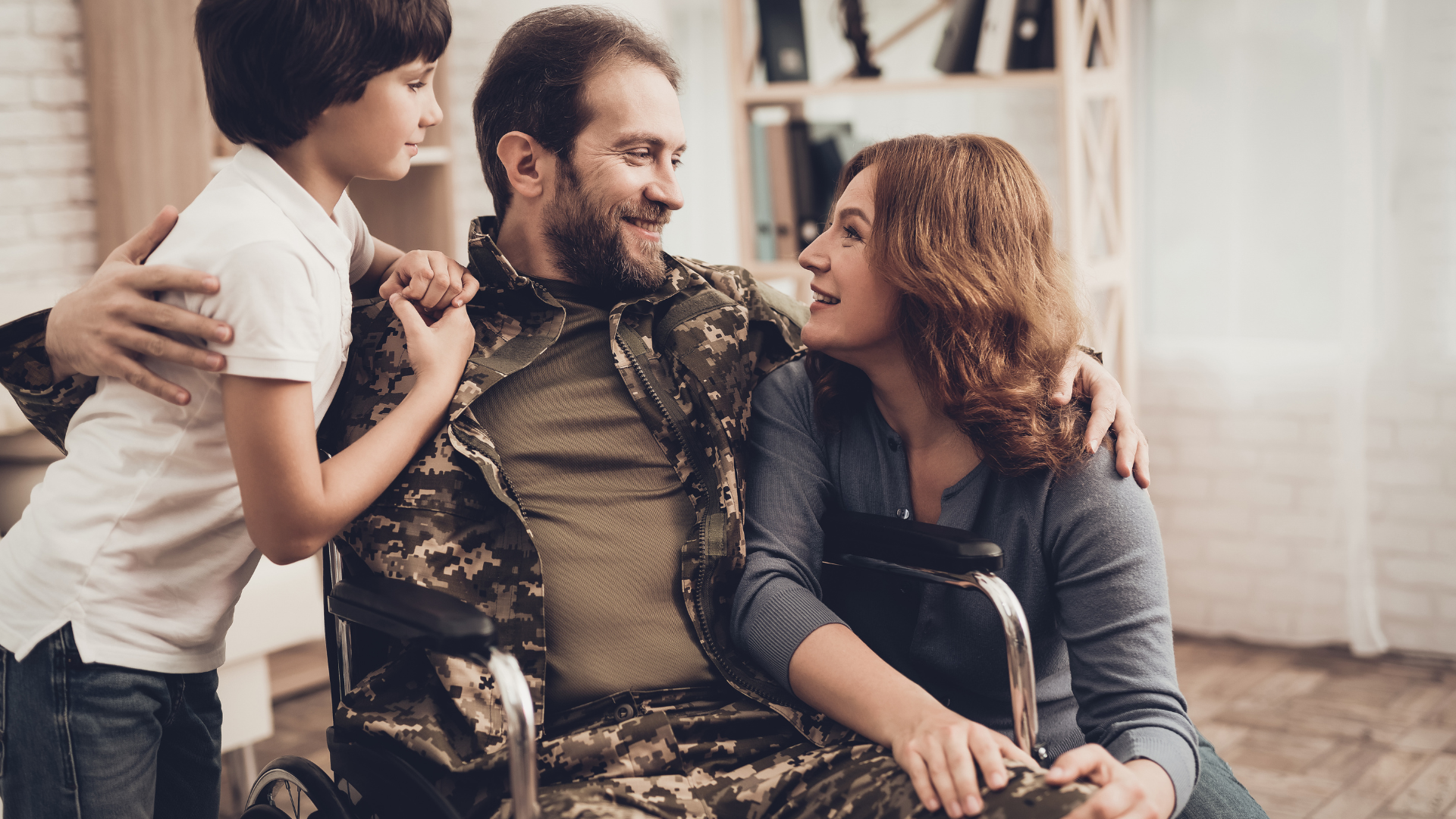 Man in military uniform in wheelchair embraced by a woman and boy indoors. Smiling, warm expressions.