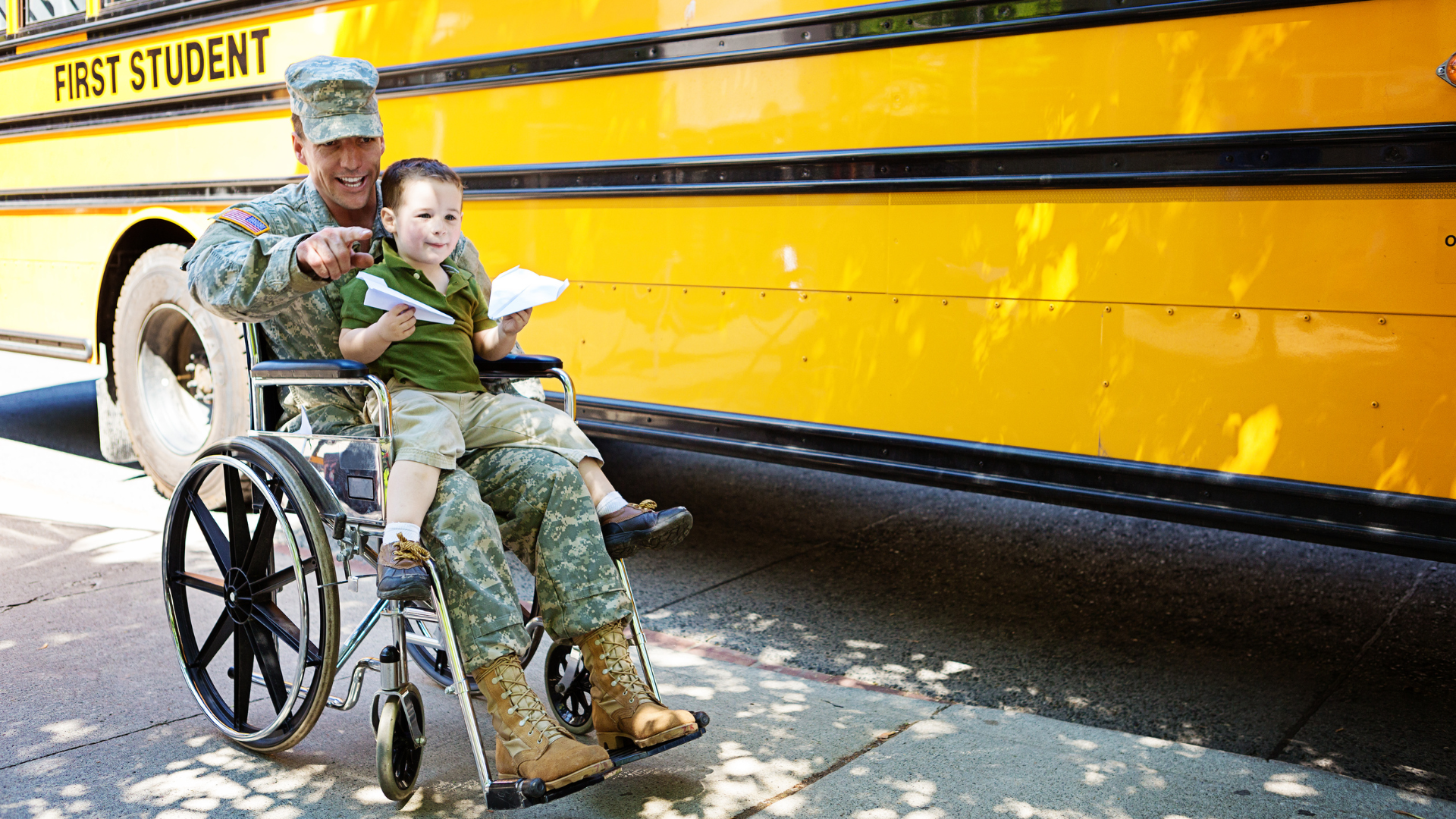 Man in military uniform holding child in his lap in a wheelchair, near a yellow school bus, smiling.