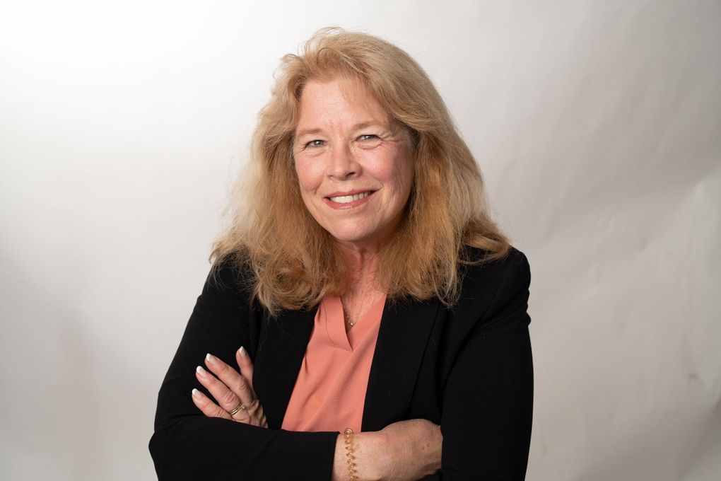 A woman in a black jacket and pink shirt is smiling with her arms crossed.