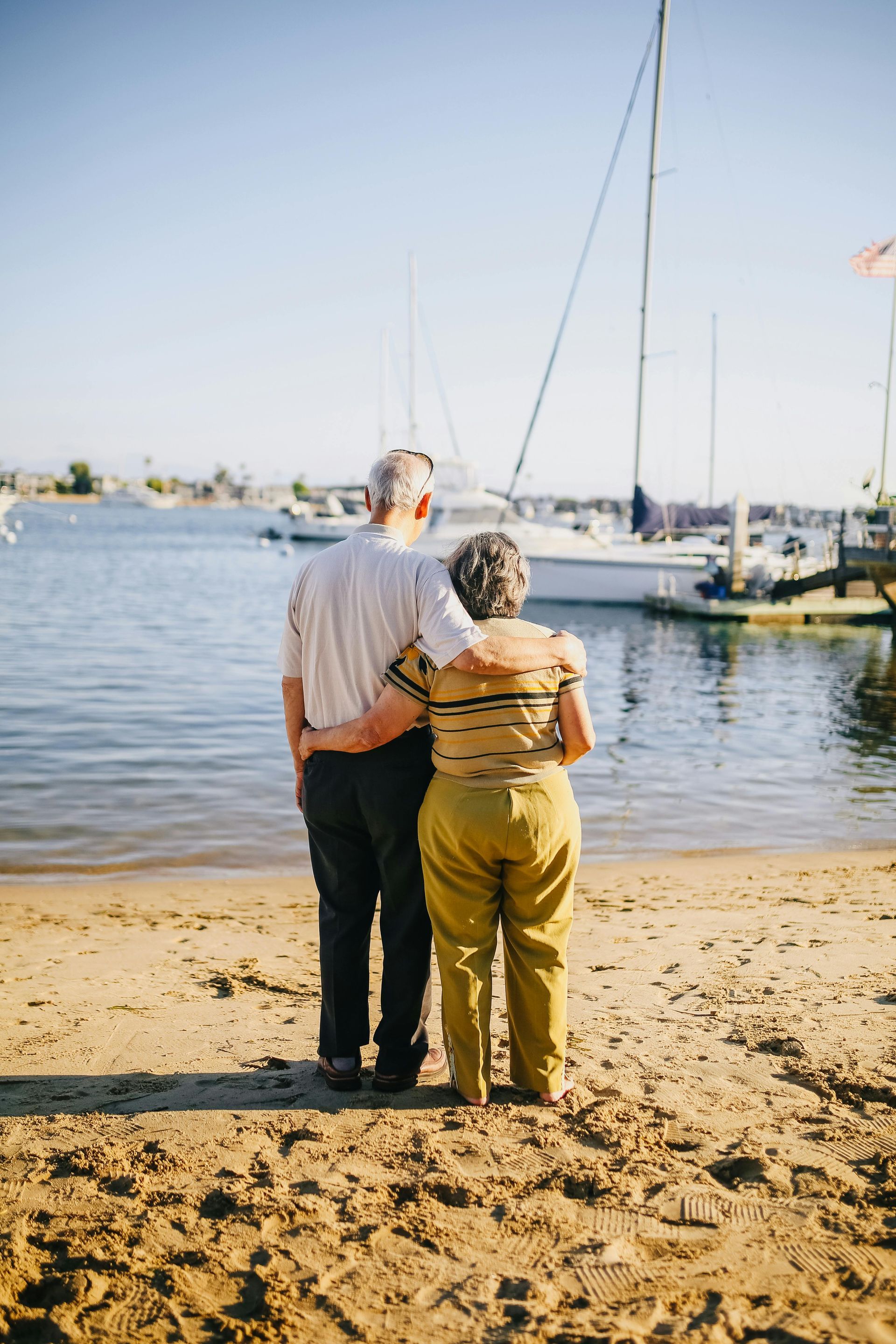 An elderly couple is standing on a beach looking at the water.