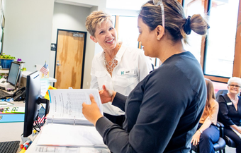 Two women at a desk in an office. One smiles as she looks at the other, who holds paperwork.