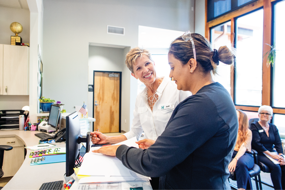 Two women smiling at a computer near a desk; one points at documents. Other people are seated nearby.