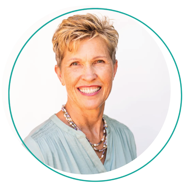 Smiling woman with short blonde hair, wearing a blue-green shirt and layered necklace, outdoors.