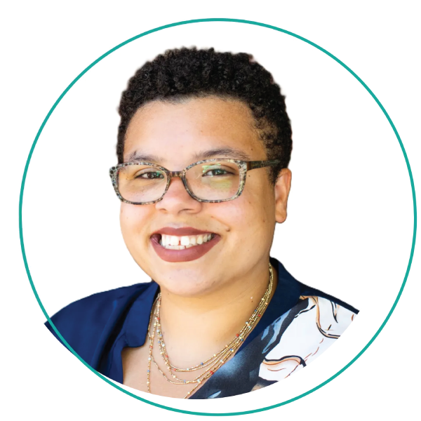 Smiling woman with short blonde hair, wearing a blue-green shirt and layered necklace, outdoors.