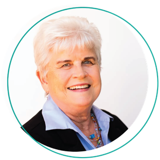 Smiling woman with short blonde hair, wearing a blue-green shirt and layered necklace, outdoors.