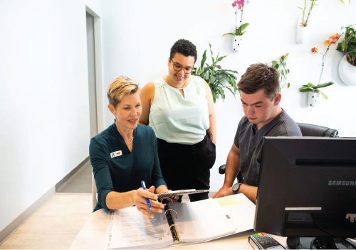 Three people at a desk review paperwork. One points with a pen while the others observe, a monitor nearby.