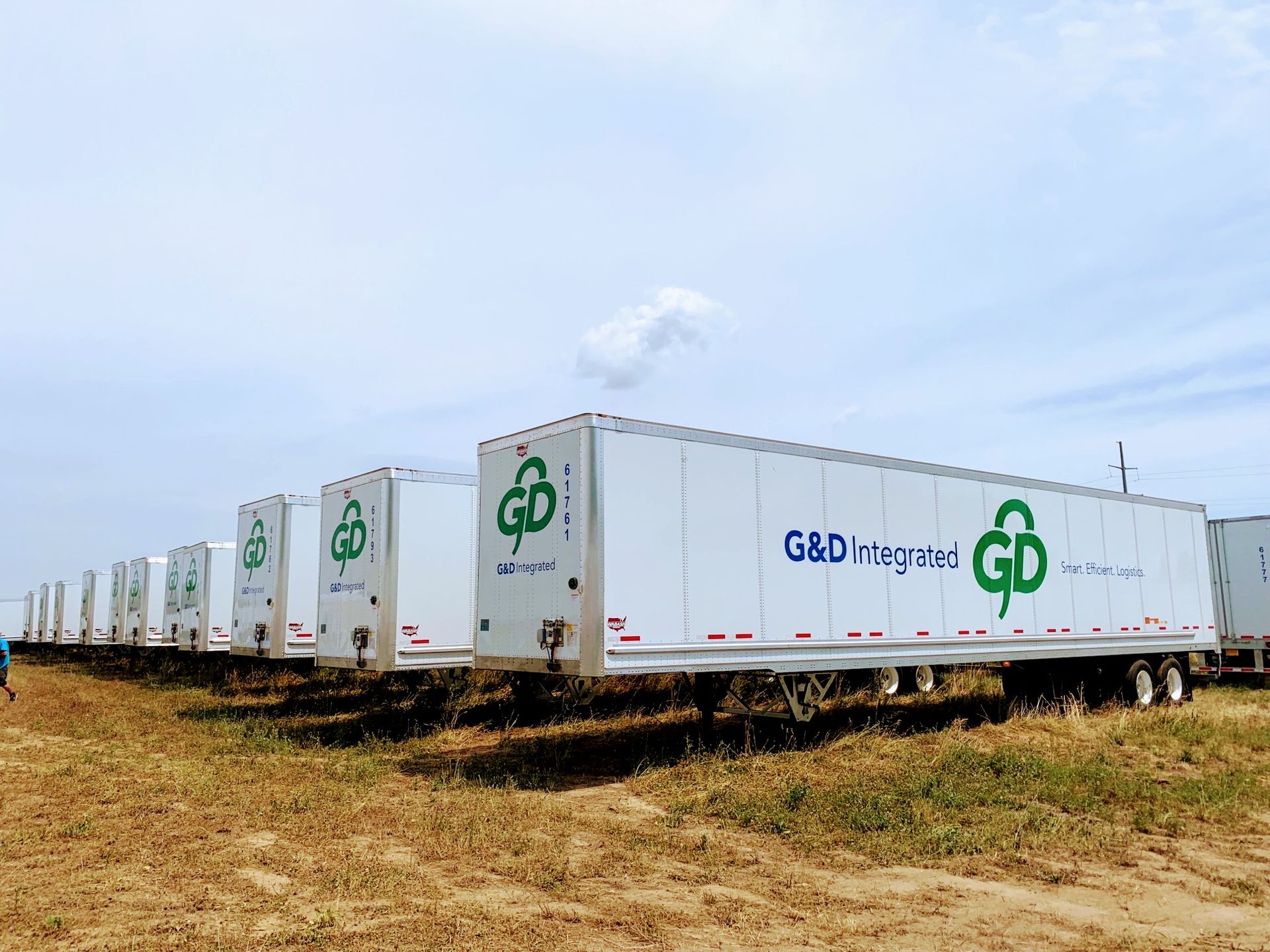 Line of white G&D Integrated semi-truck trailers parked on a field under a cloudy sky.