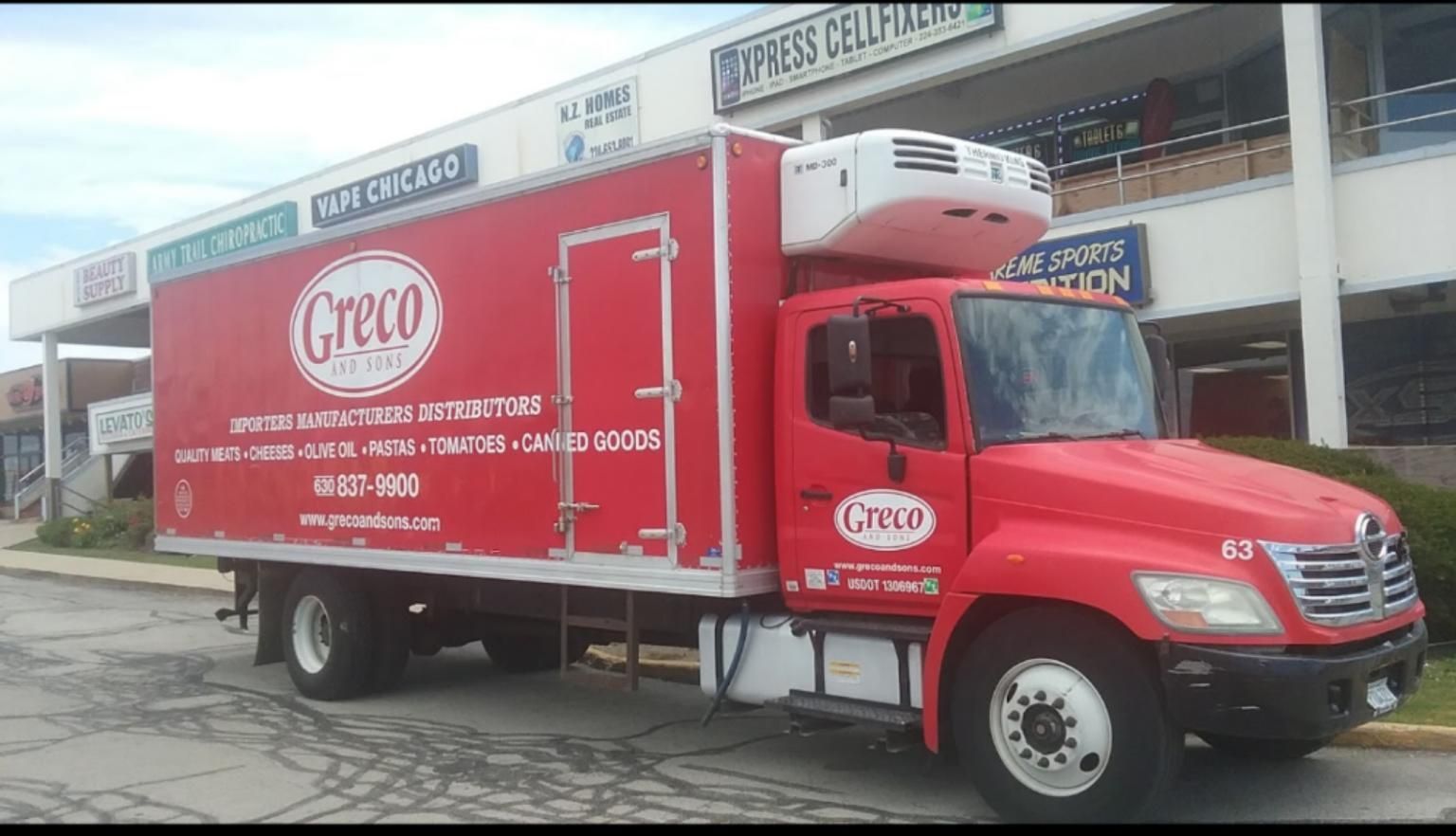 Red Greco delivery truck parked in front of a strip mall with business signs.