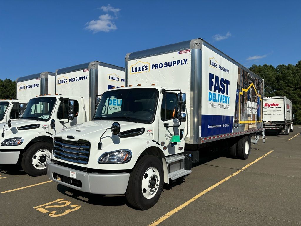 Row of red semi-trucks parked on asphalt under a blue sky, some with logo on trailer.