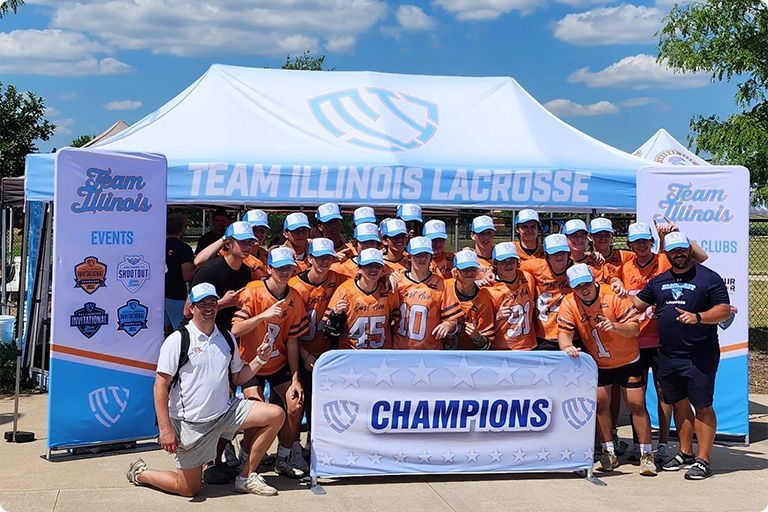 Team Illinois Lacrosse team in orange jerseys celebrate championship win. Tent backdrop reads 