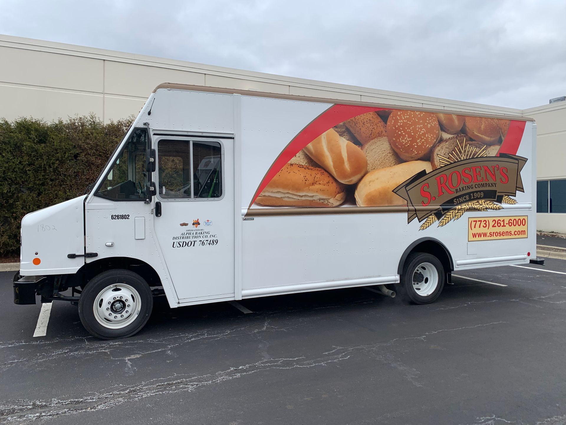 White bakery delivery truck with bread graphic on the side, parked in a lot.