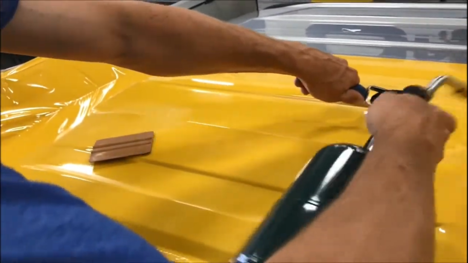 Person using a heat gun on yellow vinyl wrapping a vehicle, wooden smoothing tool nearby.