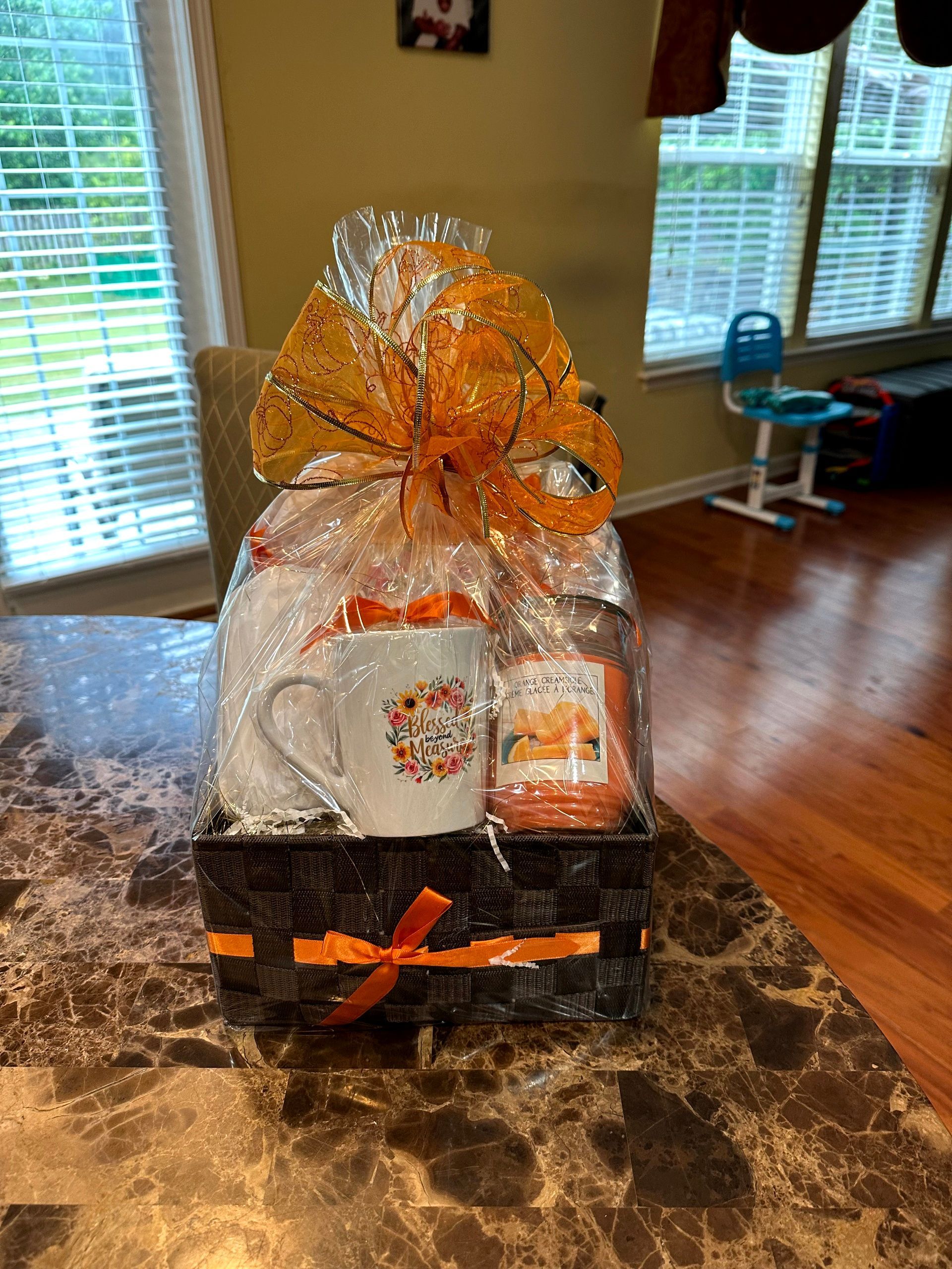 Gift basket with fall-themed mug, candle, and orange ribbon, on a table near a window.