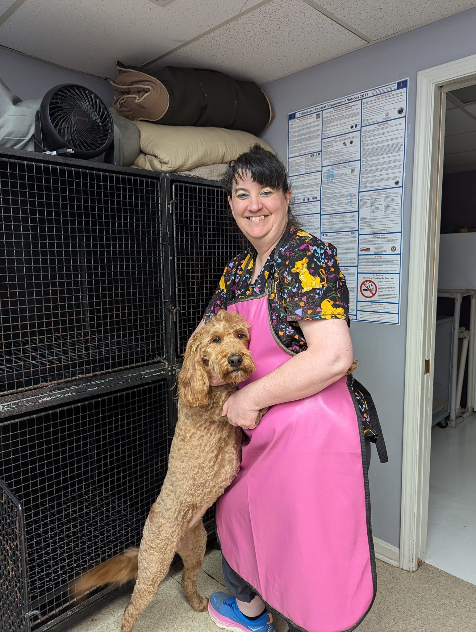 A woman in a pink apron is holding a dog in front of a cage.