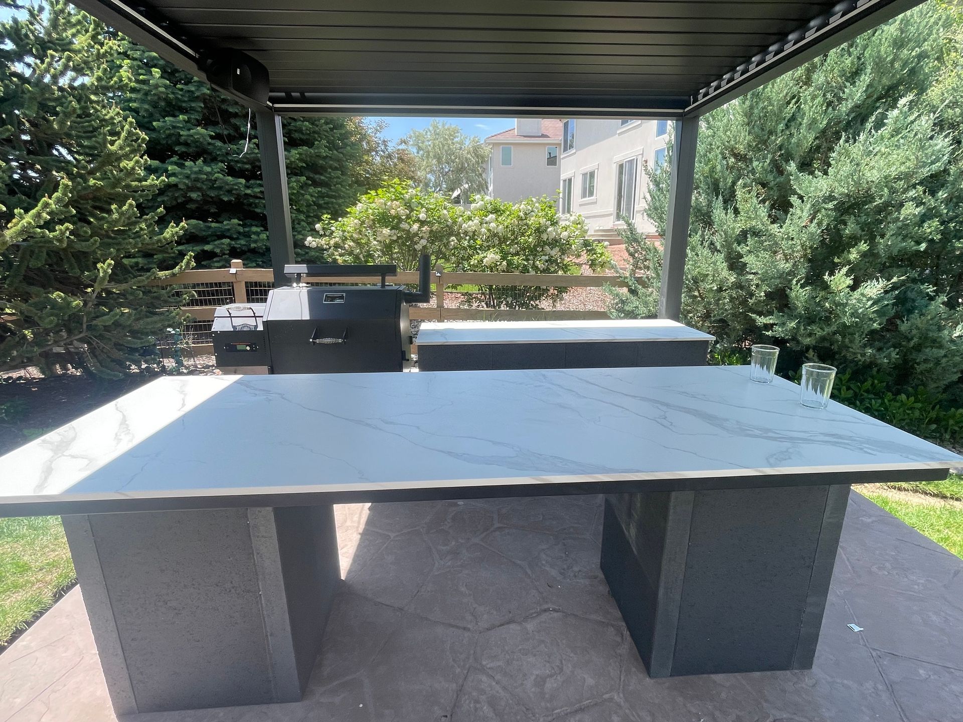 A large table with a marble top is sitting under a pergola.