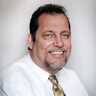 A man with a beard is wearing a white shirt and tie and smiling.