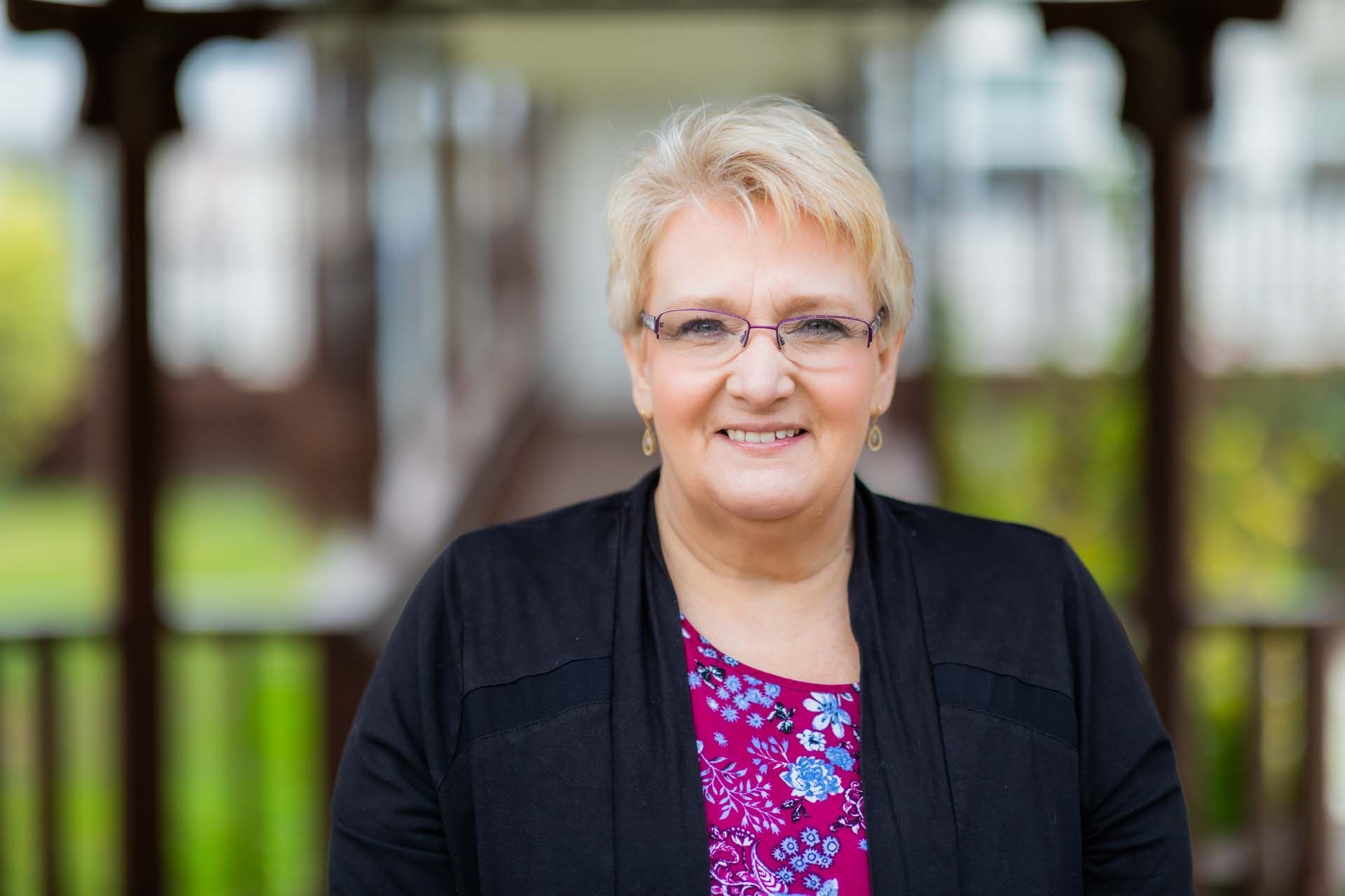 A woman wearing glasses and a black jacket is standing in front of a gazebo.