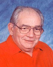 Jimmy Simpson wearing glasses and a red shirt is smiling for the camera.