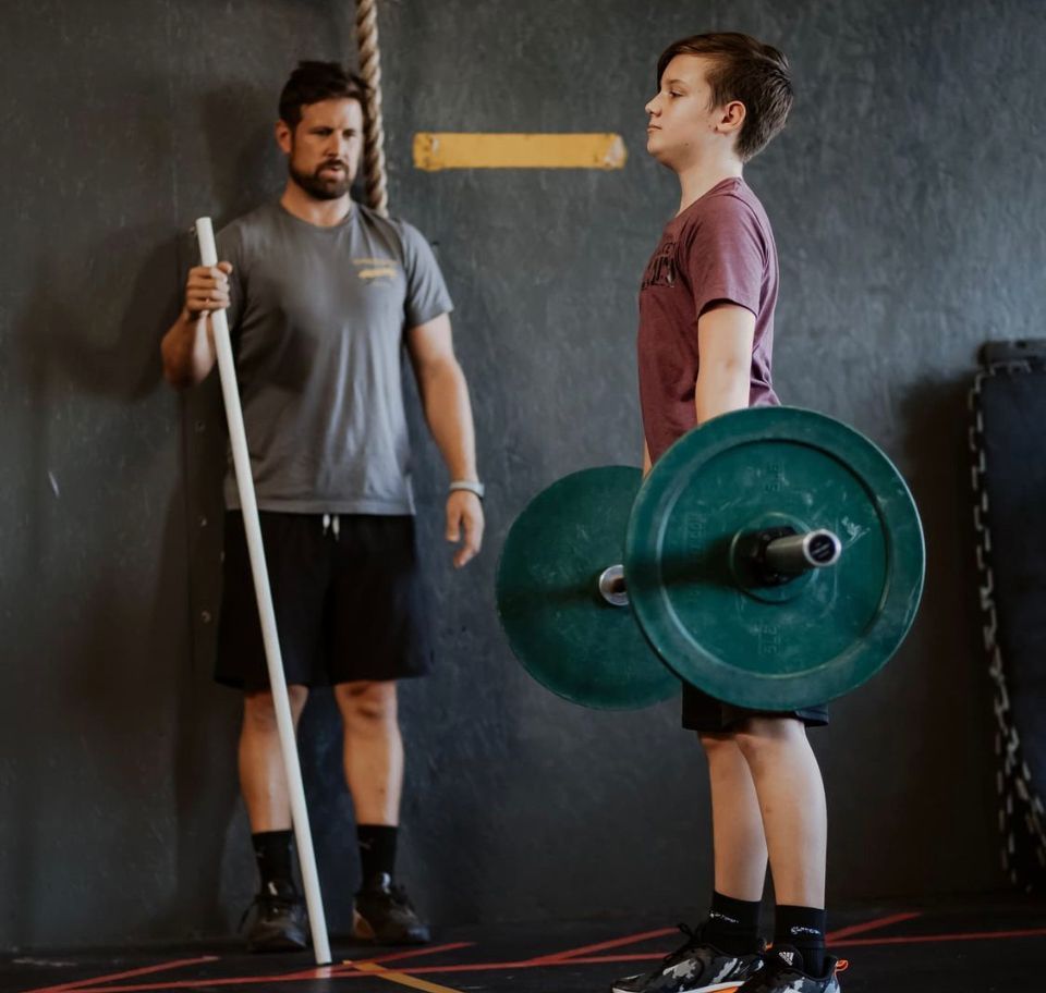 Two athletes in a gym, one holding a barbell and another standing with a training staff nearby.