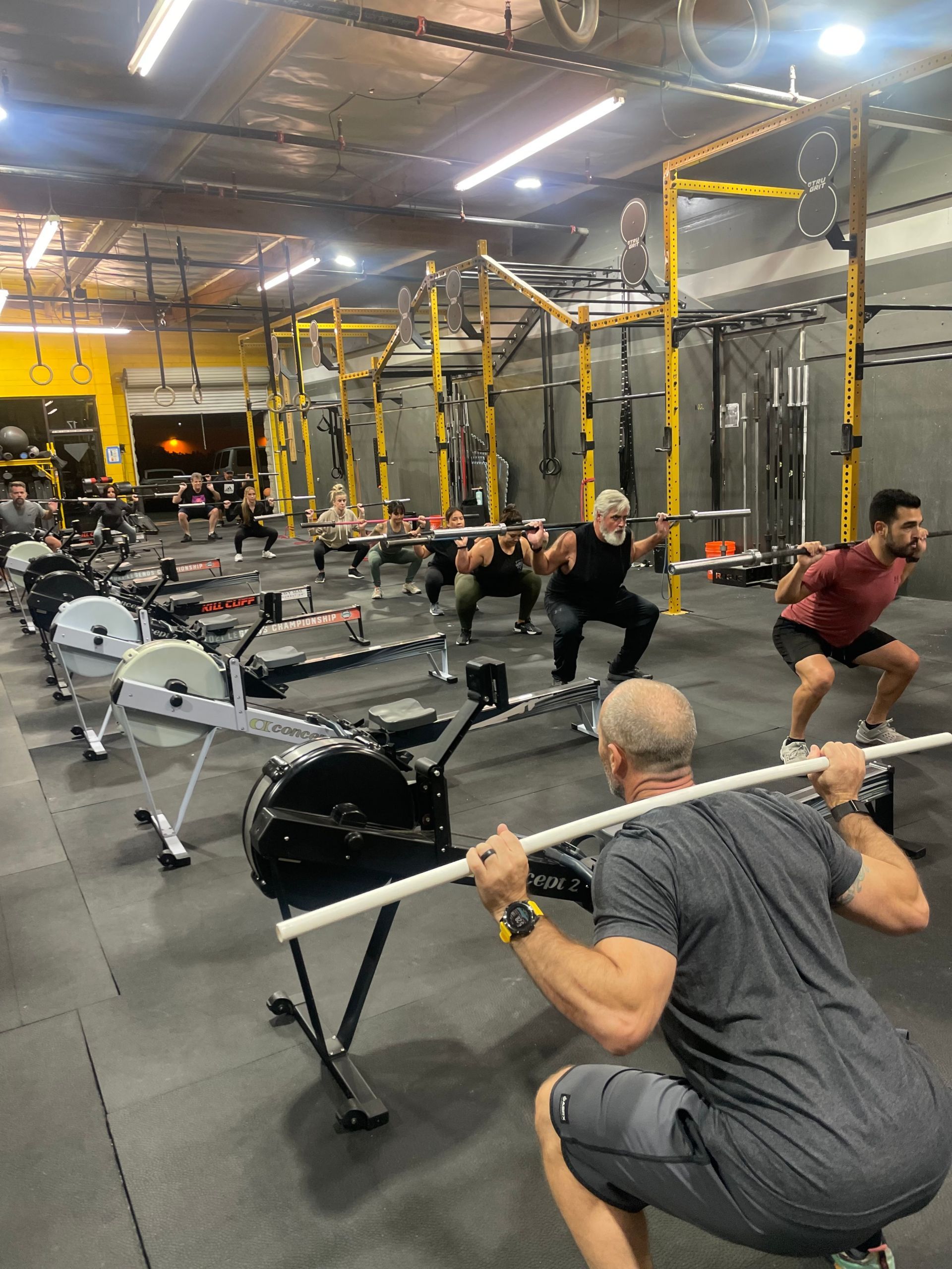 People doing barbell squats in a gym with yellow racks and rows of equipment