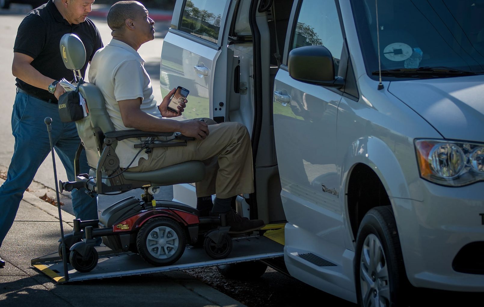 A person is being assisted into a black car by a person in a black shirt, near a modern building.