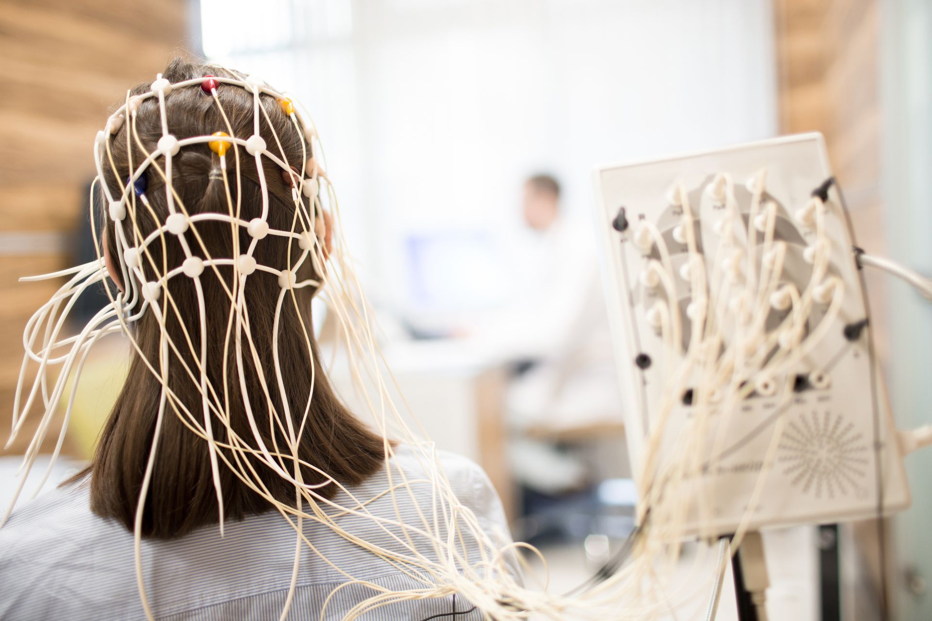 Woman wearing a white EEG cap in a medical setting. Wires connect to a monitoring device, researcher in background.
