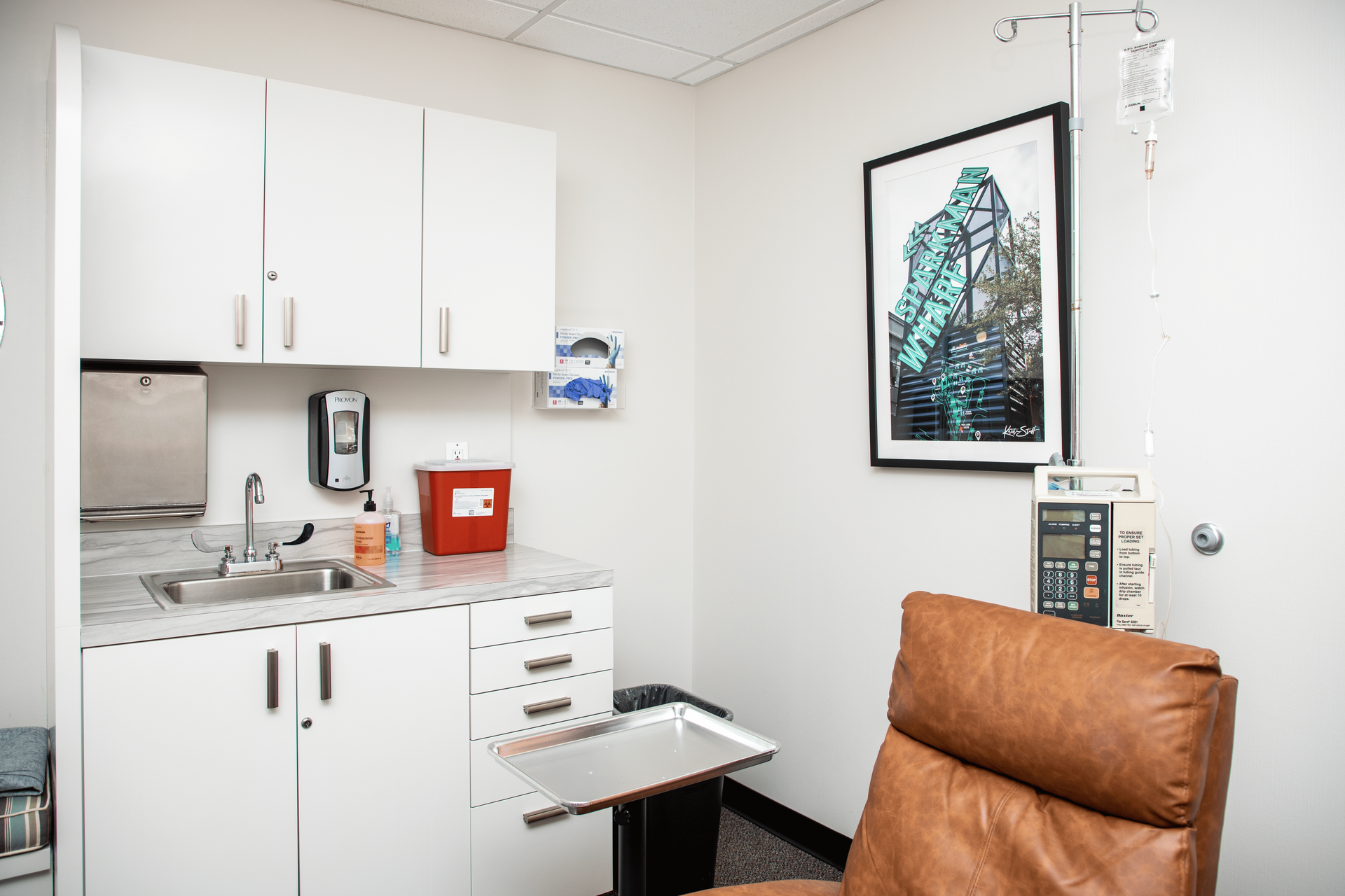A doctor 's office with a chair , sink , cabinets and a picture on the wall.
