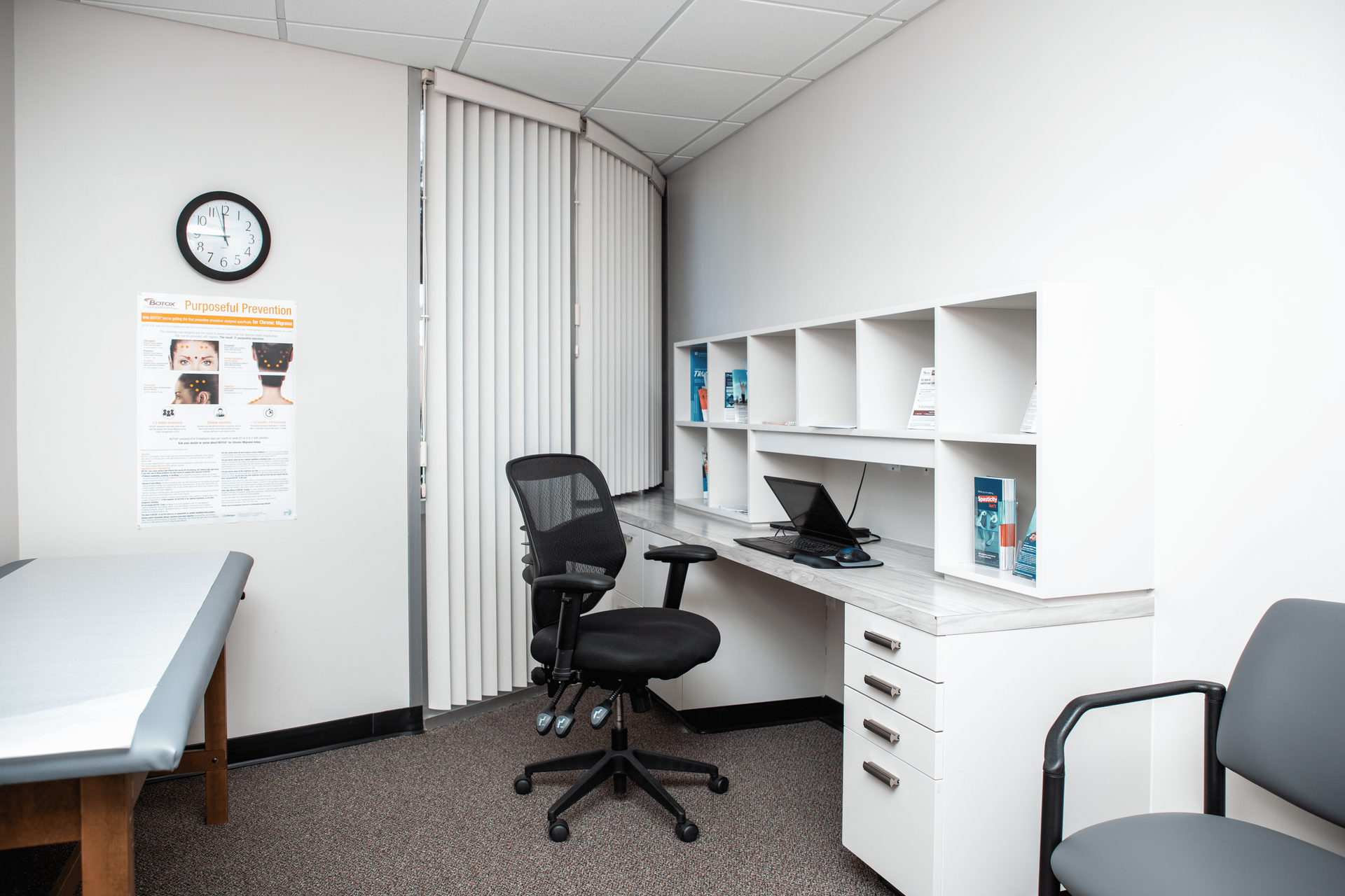 A doctor 's office with a desk , chair , and a clock on the wall.