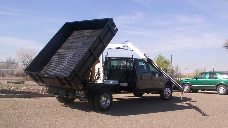 Brown dump truck with raised bed, parked on gravel. A green SUV is in the background.