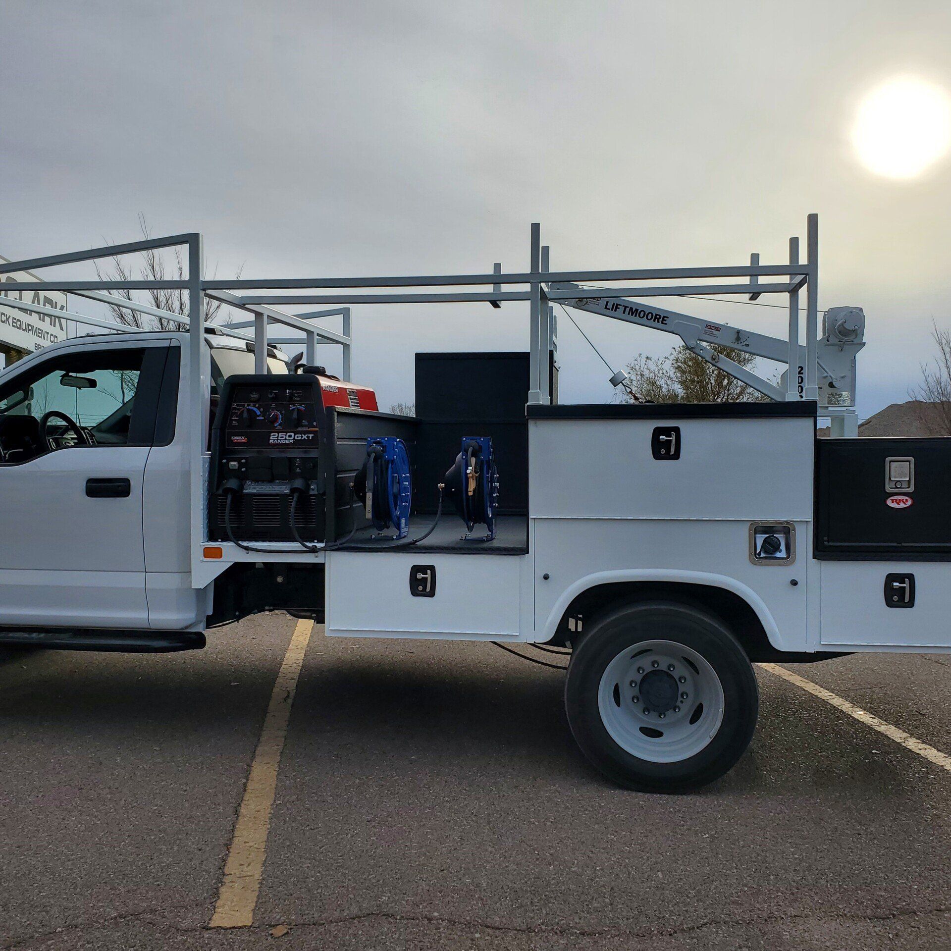 White work truck with a crane and tool compartments, parked outdoors on a cloudy day.