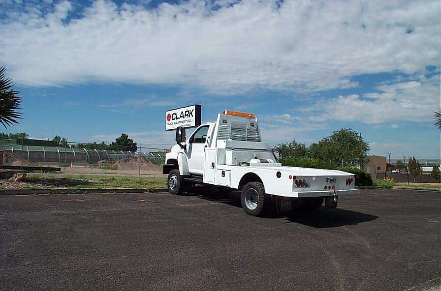 White flatbed truck parked in a lot under a blue sky with clouds. The truck has a service body and a sign.
