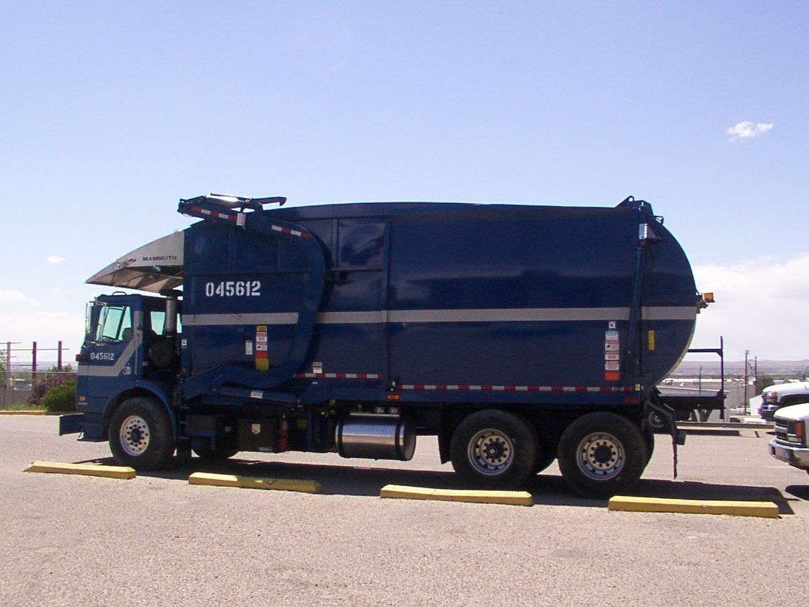 Blue garbage truck parked on gravel under a sunny sky.