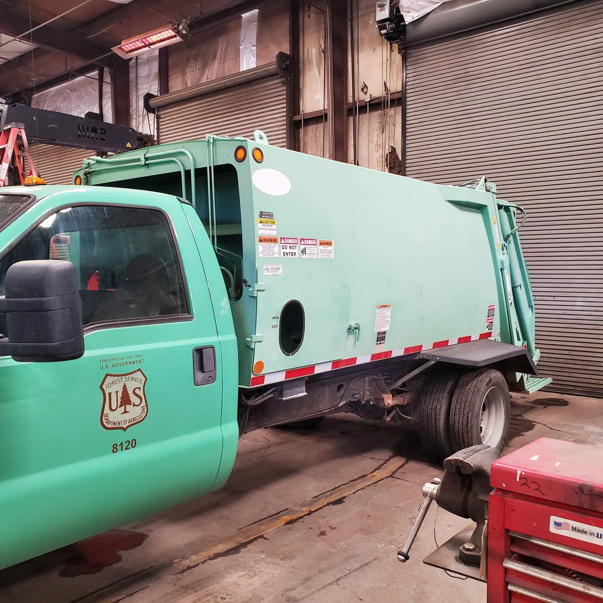 A green garbage truck parked in a garage. The truck has a US Forest Service logo on the door.