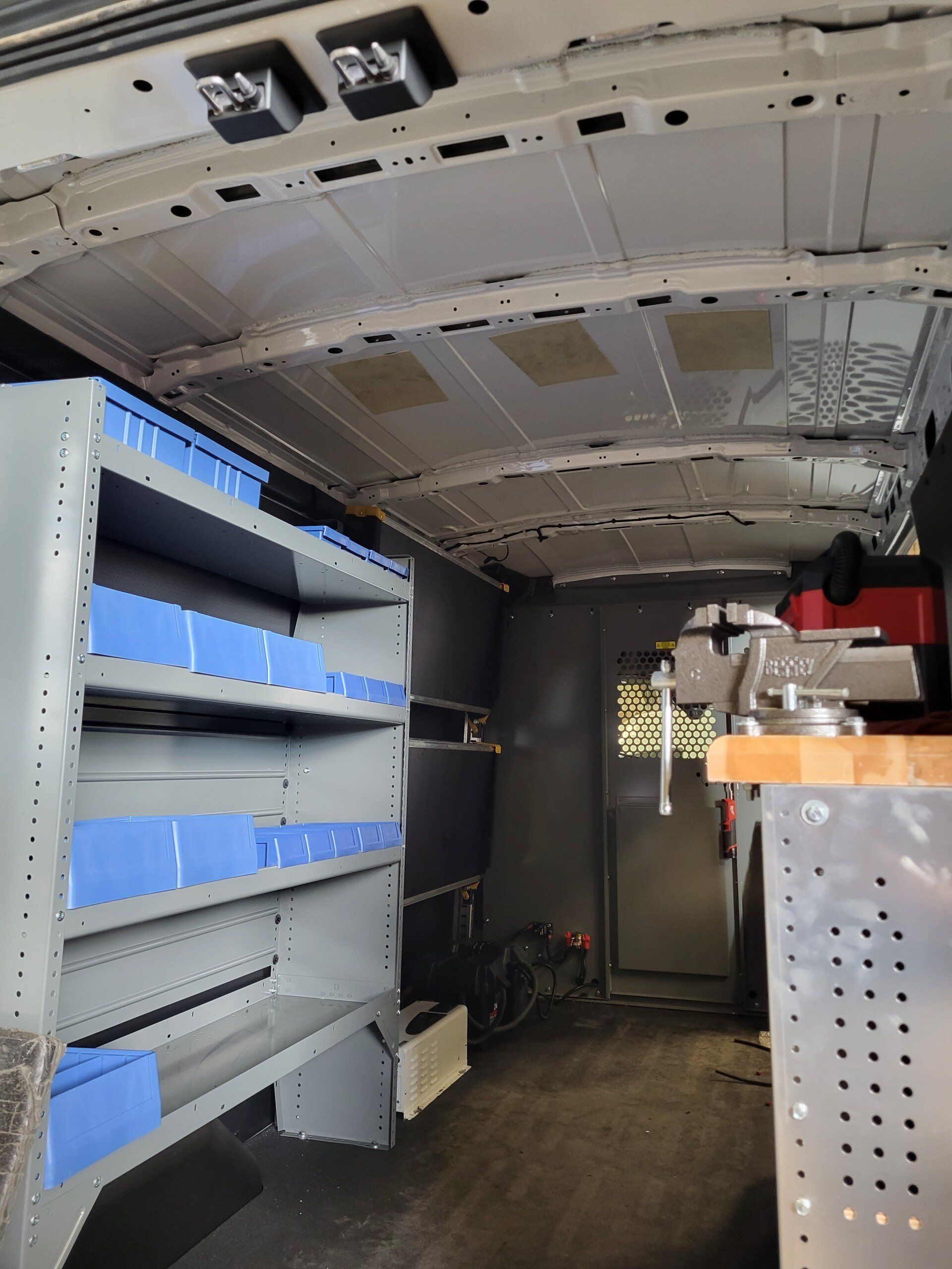 Interior of a van, with shelving, blue bins, and a workbench. Grey and blue color scheme.