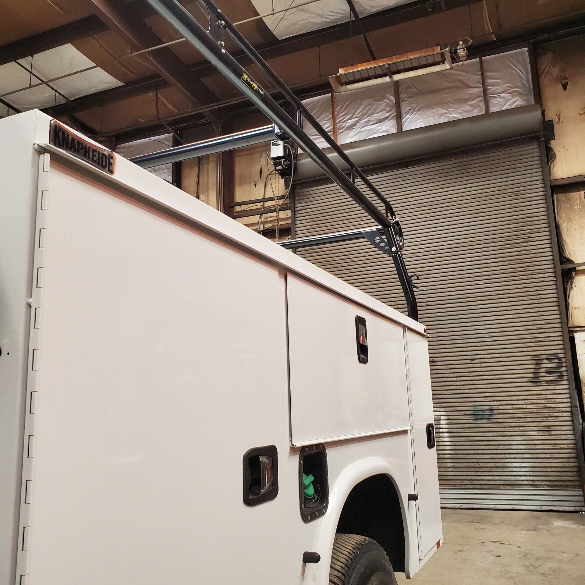 White utility truck with a ladder rack in a warehouse, next to a closed gray roll-up door.