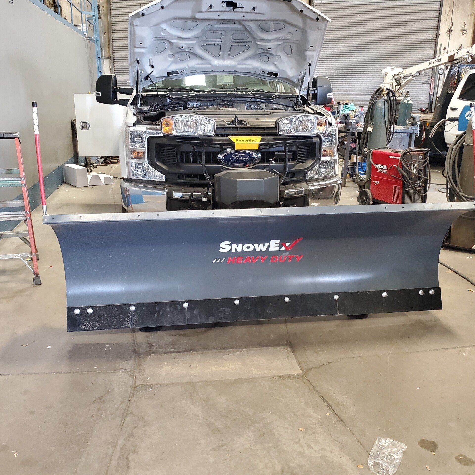 Snow plow attached to a white truck inside a repair shop.