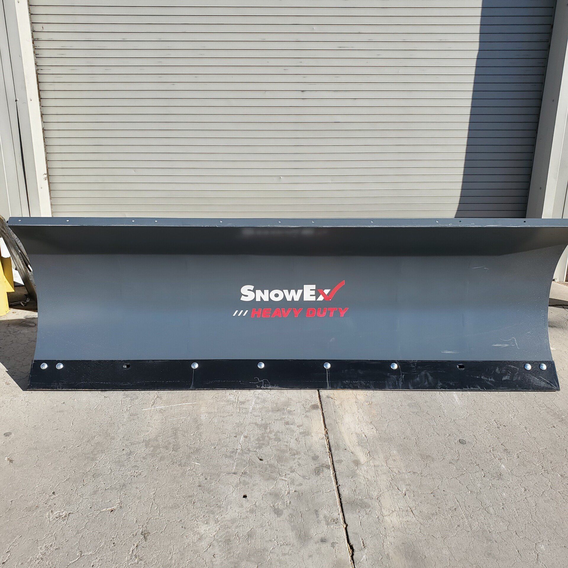 SnowEx heavy-duty snowplow. Gray blade with black rubber edge. The setting is outdoors, in front of a garage door.