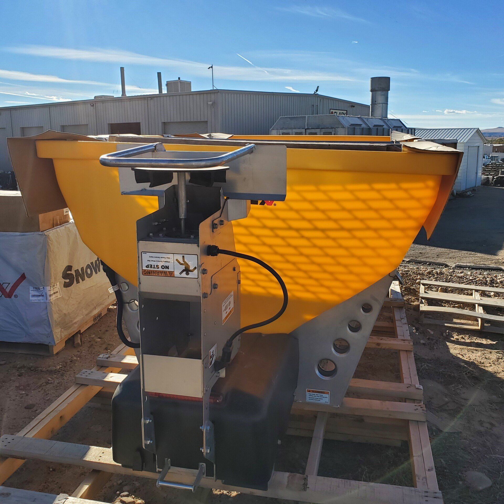 Yellow salt spreader on a wooden pallet outdoors, against a building, clear sky.