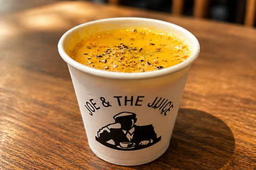 Small paper cup of soup with black pepper on a wooden table, labeled “Joe & the Juice”