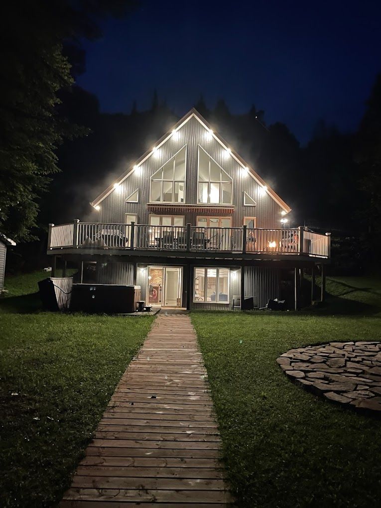 Une grande maison est éclairée la nuit avec une passerelle en bois qui y mène.