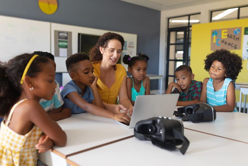 A teacher is teaching a group of children how to use a laptop computer.