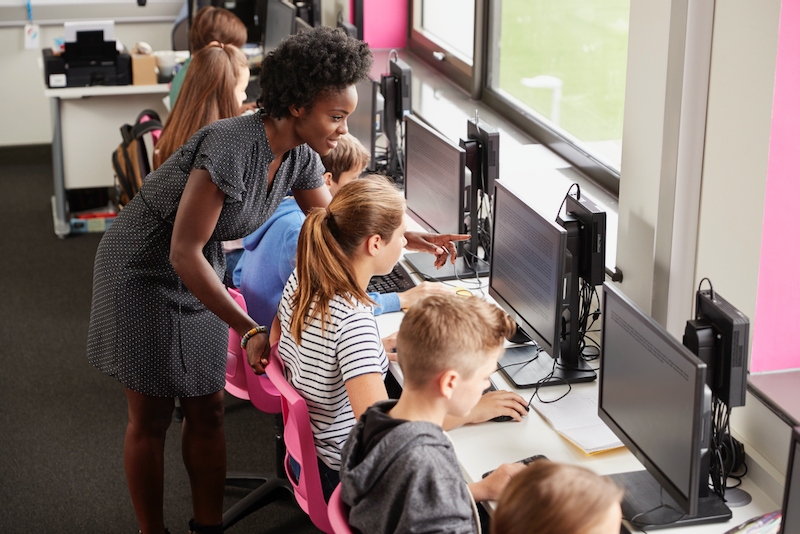 A teacher is helping a group of children use computers in a classroom.