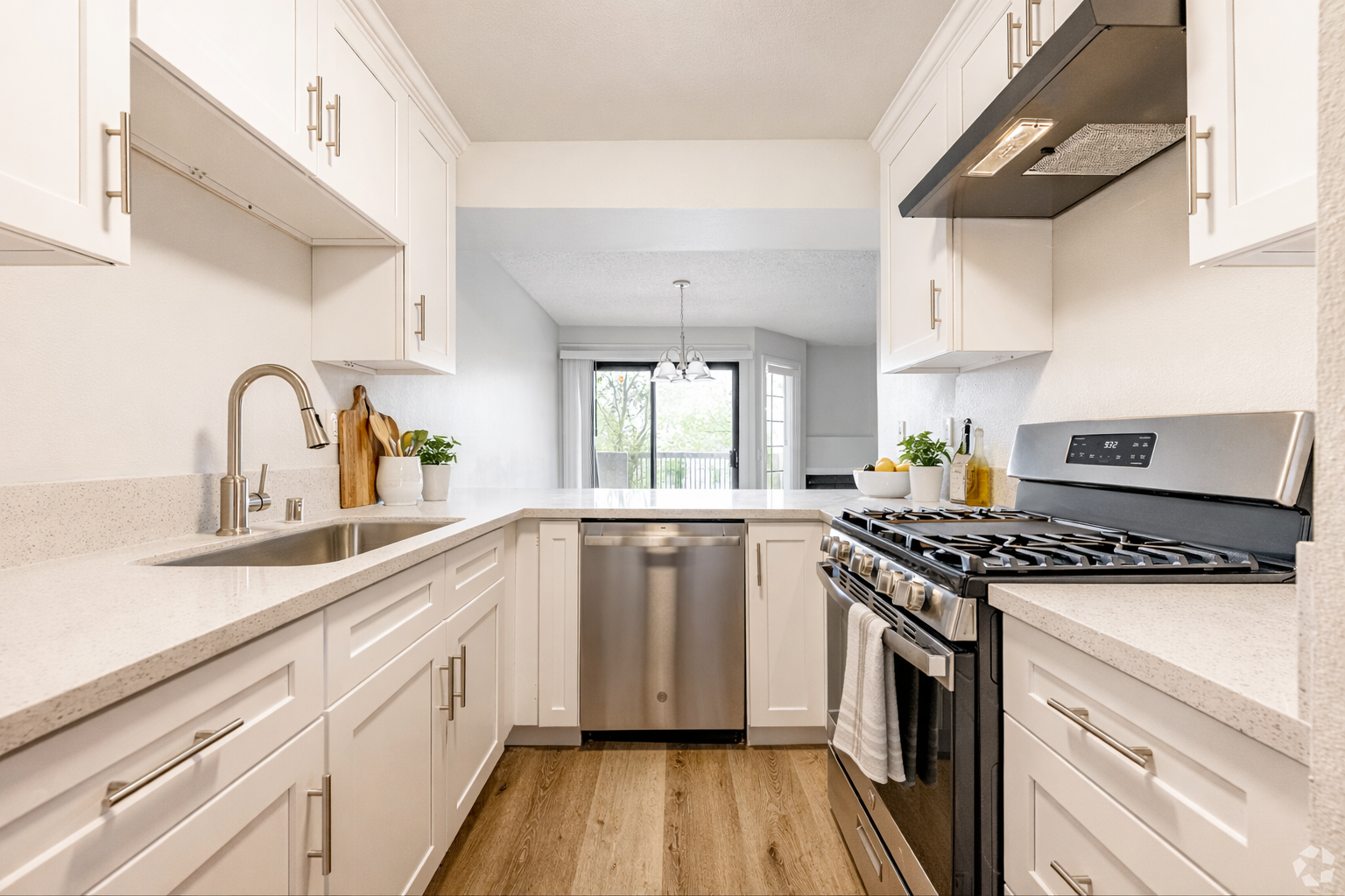 A small, bright white kitchen with a stainless steel range and dishwasher. View into a living area.