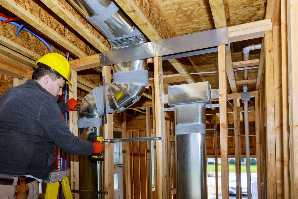A person wearing a yellow hard hat and orange gloves installs metal ductwork in an unfinished wooden building frame.