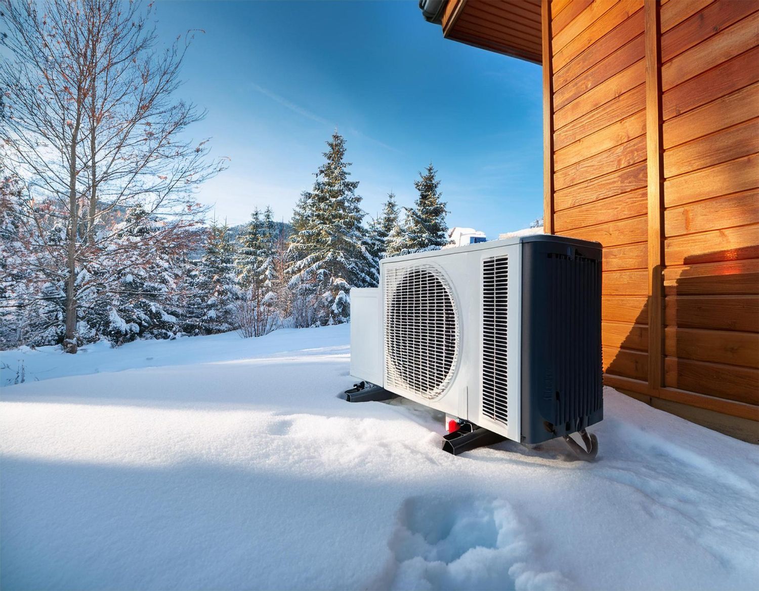 Heat pump unit in snow next to a wooden building on a sunny winter day.